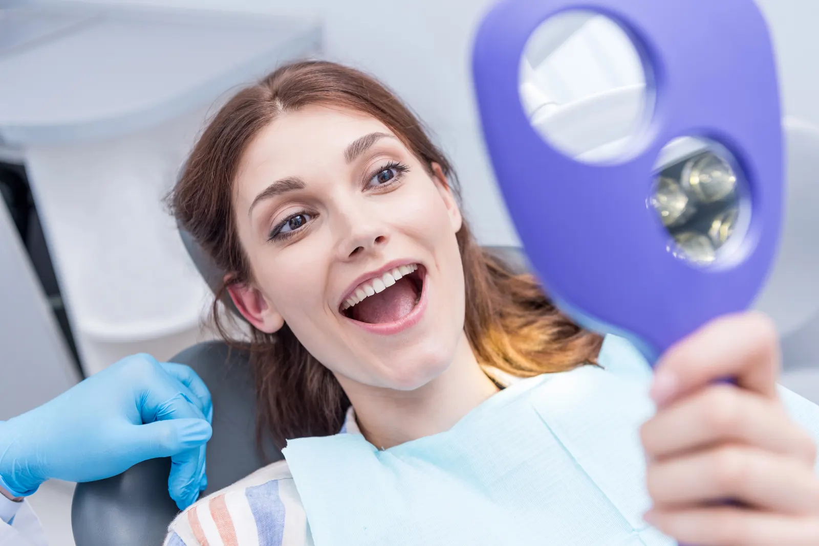 Happy woman in dental chair holding a mirror and smiling, showing her teeth after dental checkup.