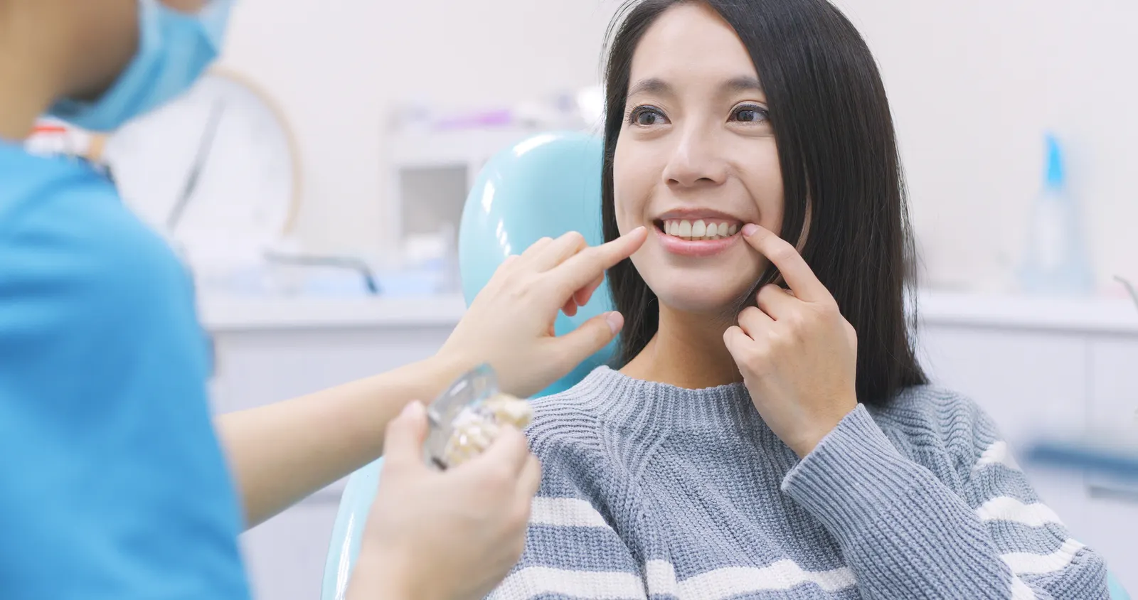 Smiling woman pointing to her teeth while dentist in blue scrubs demonstrates dental model.