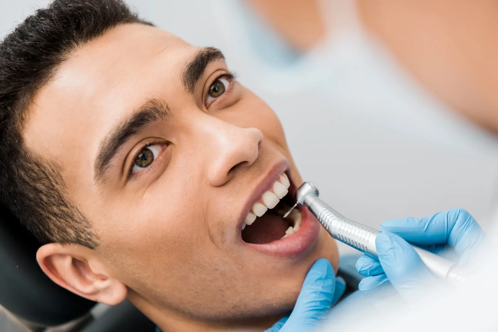 Dentist wearing blue gloves cleaning a young man's teeth with a dental drill.