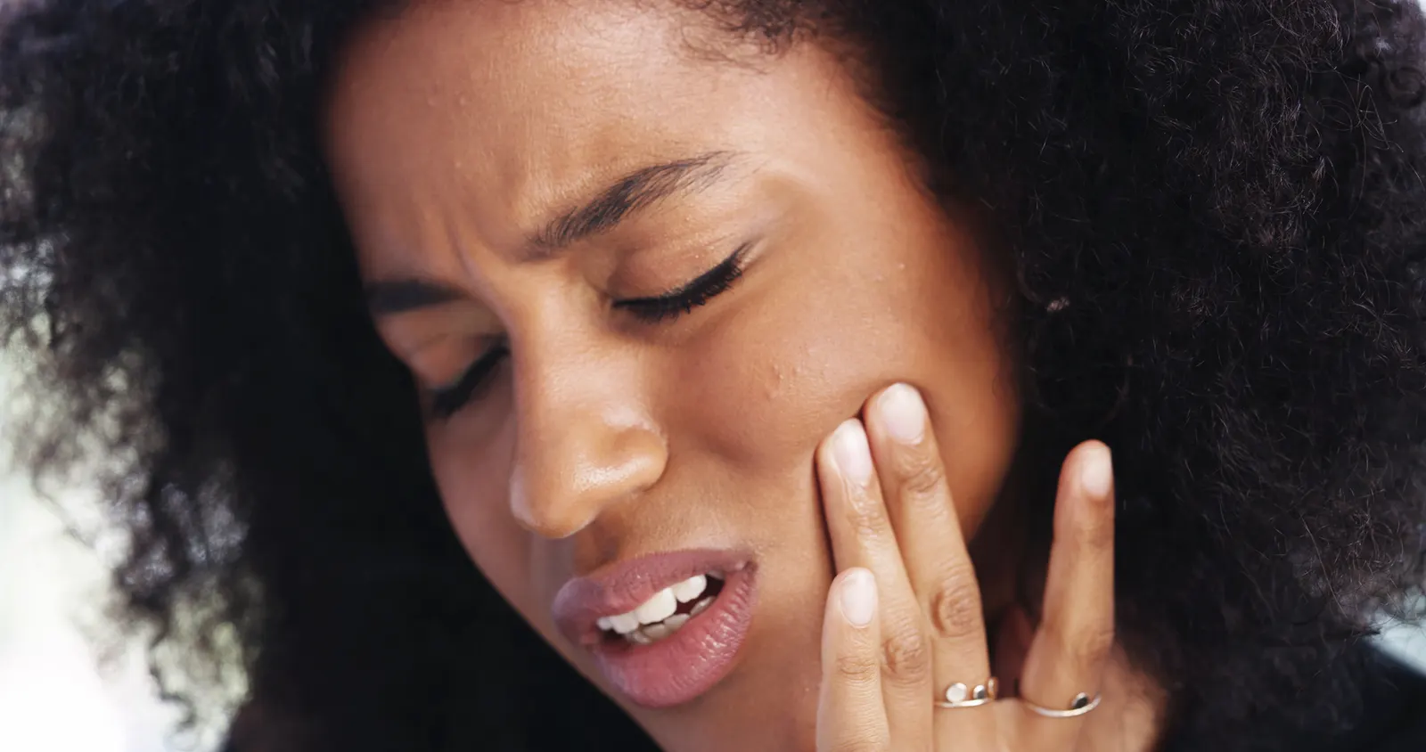 Woman with curly hair holding her cheek, showing a pained expression, indicating toothache or pain.