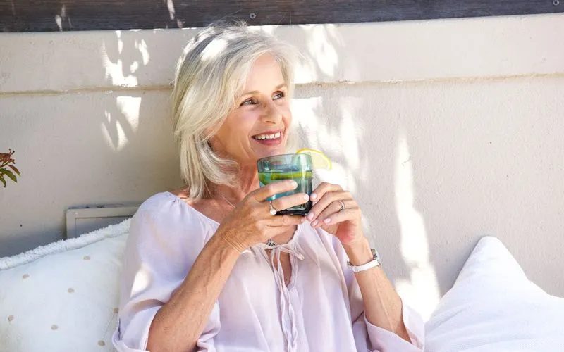Smiling older woman with gray hair sitting outdoors, holding a glass with a lemon slice.