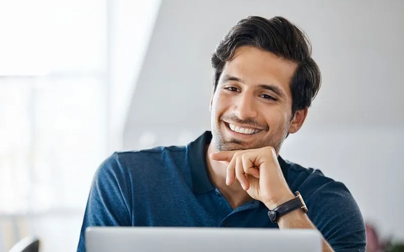 Smiling man with dark hair wearing a blue polo shirt, sitting and looking at a laptop screen.