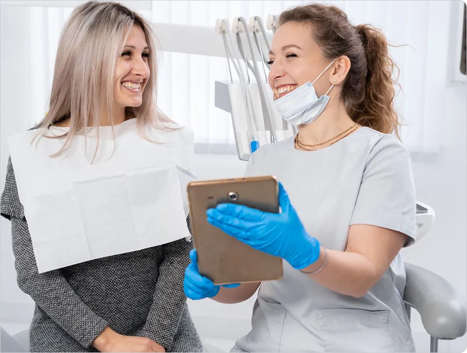 Dentist with gloves and mask smiling, showing tablet to smiling patient in dental clinic.